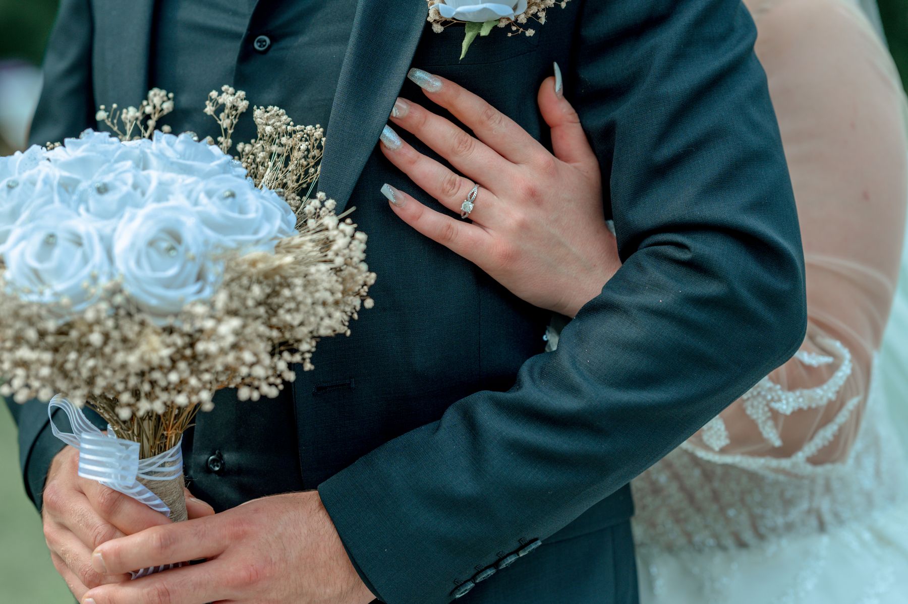 Wedding couple embracing beside a white rose bouquet, with a hand and ring resting on a dark suit.